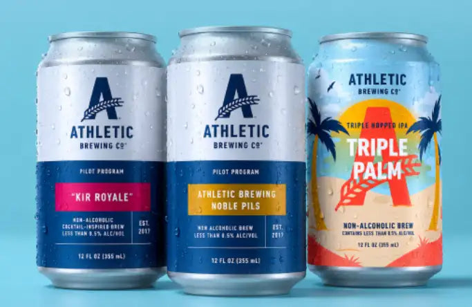 Three aluminum beverage cans from athletic brewing co., featuring distinct designs: one with a blue and white label for ’kir royale,’ another in navy blue for ’athletic brewing noble pils,’ and the third brightly colored with palm trees and orange accents labeled